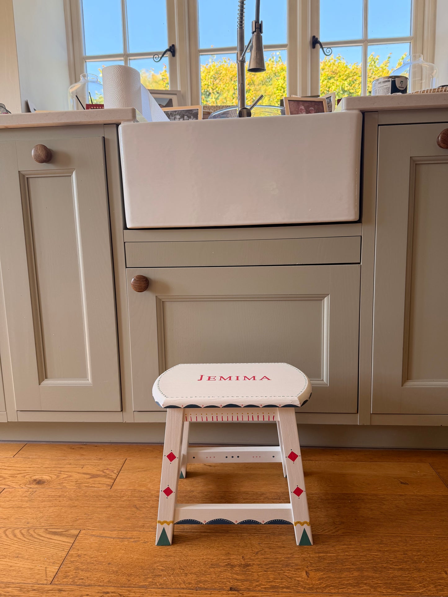 A white wooden step stool with the name 'JEMIMA' printed on the backrest, positioned next to a light gray kitchen cabinet in a home setting with hardwood floors.