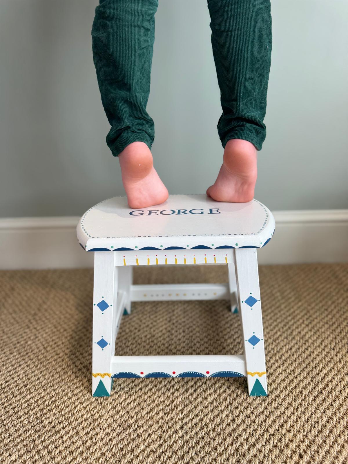 Child's feet on a small step stool with 'GEORGE' on the seat, on a carpeted floor.