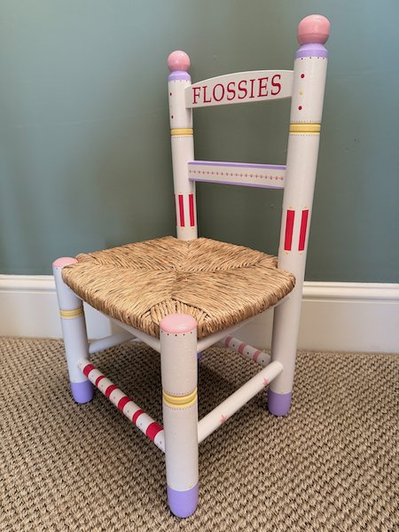 A whimsical children's chair with a woven straw seat, painted white frame, pink and purple accents, and the word 'FLOSSIES' on the backrest, set on a carpeted floor against a blue wall.