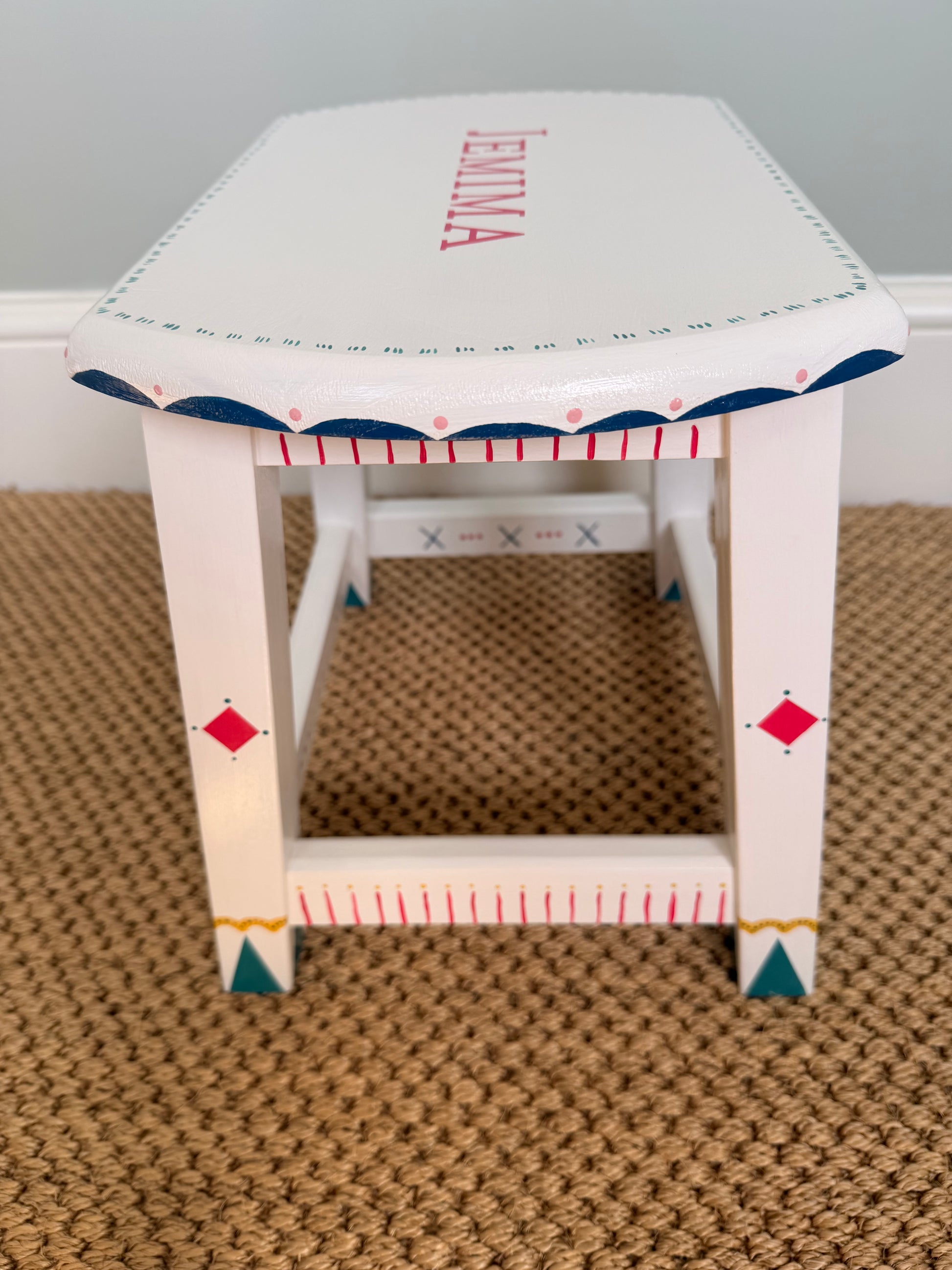 A white wooden stool with personalized pink lettering reading 'JEMIMA' on the side, decorated with blue scalloped trim, red measurement markings, and small geometric accents, shown on a textured tan carpet.