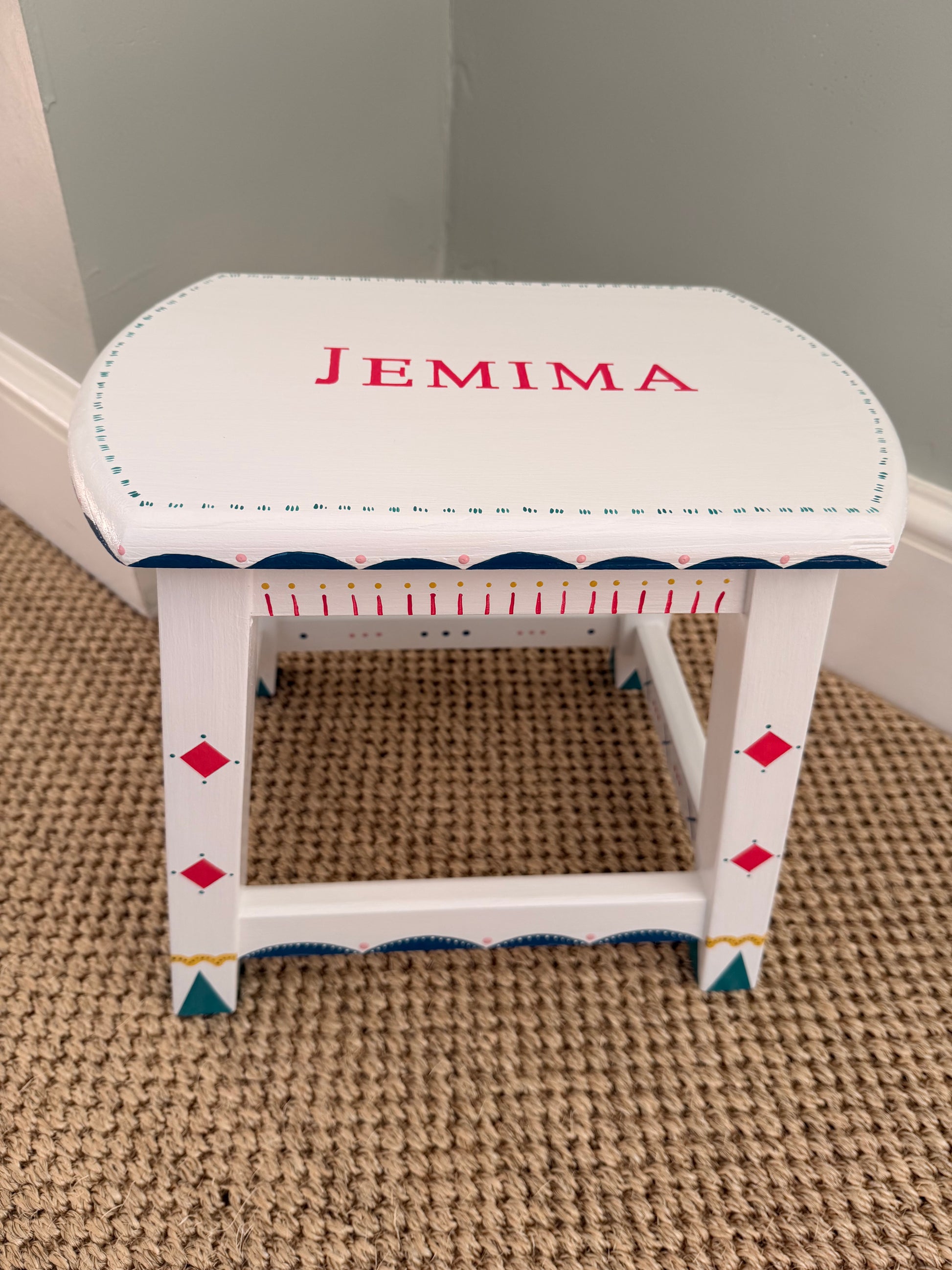 A white wooden stool with decorative red, blue, and yellow painted details, personalized with the name 'JEMIMA' in red on the seat, set on a beige carpeted floor.