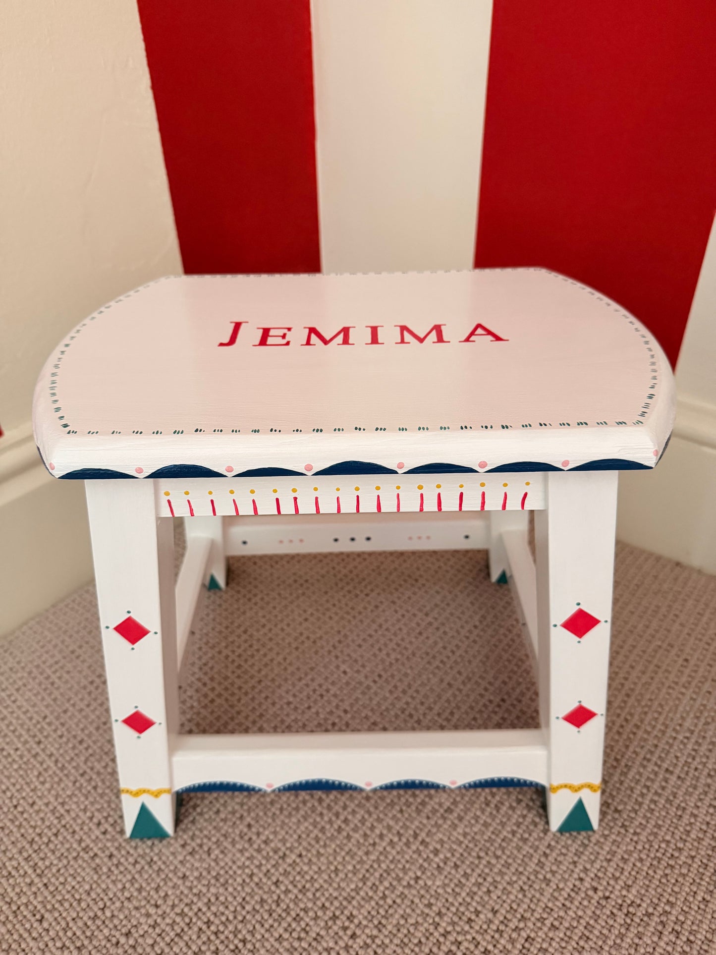 A white wooden stool with a curved backrest, personalised with the name 'JEMIMA' in red. The sides feature blue and red decorative details, including scalloped edges and diamond motifs, set against a red and white striped wall and a beige carpet.