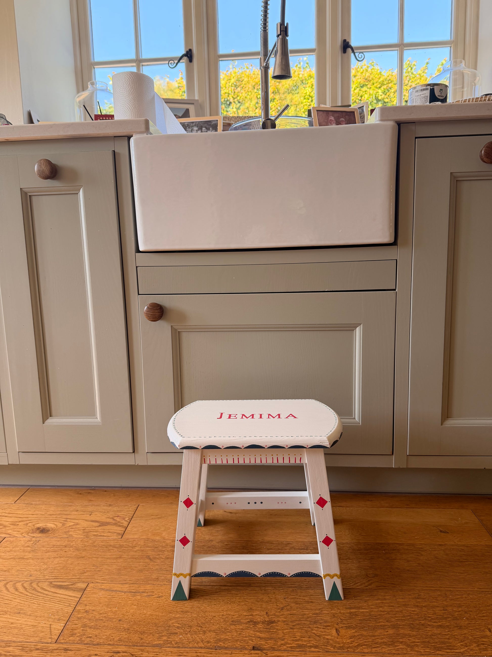 A white wooden step stool with the name 'JEMIMA' printed on the backrest, positioned next to a light gray kitchen cabinet in a home setting with hardwood floors.
