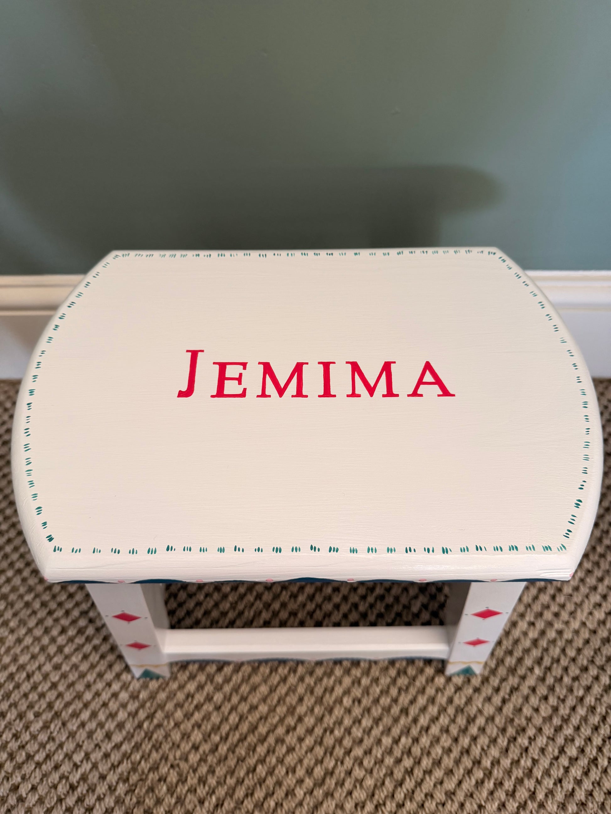 A small white wooden stool or bench with the name 'JEMIMA' painted in pink on the top, decorated with blue dotted borders and small red diamond accents on the legs, set on a carpeted floor against a painted wall.
