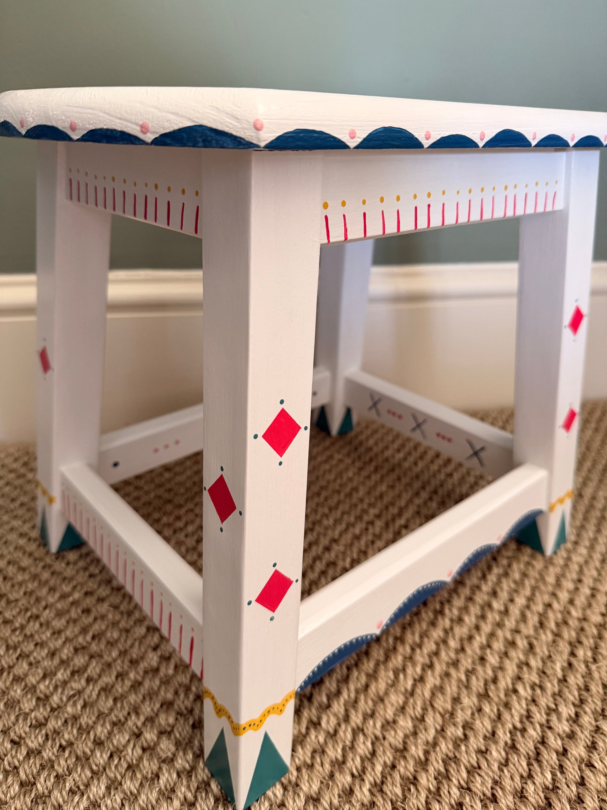 A white wooden stool with decorative geometric patterns in red, blue, green, and gold, shown on a beige carpeted floor.