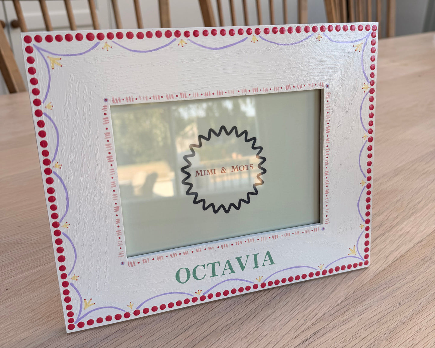 A square white picture frame with a red dotted border and decorative scrollwork, placed on a light wood tabletop. The frame has a central cutout with a black scalloped edge and the word 'OCTAVIA' printed vertically on the right side.