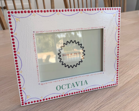 A square white picture frame with a red dotted border and decorative scrollwork, placed on a light wood tabletop. The frame has a central cutout with a black scalloped edge and the word 'OCTAVIA' printed vertically on the right side.