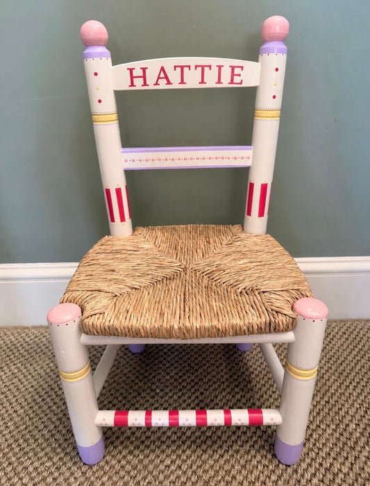 A small painted wooden chair with a woven straw seat, personalized with the name 'HATTIE' in red on the backrest. The chair features white-painted legs and arms with pink, purple, and gold decorative accents, set against a blue-gray wall and carpeted floor.