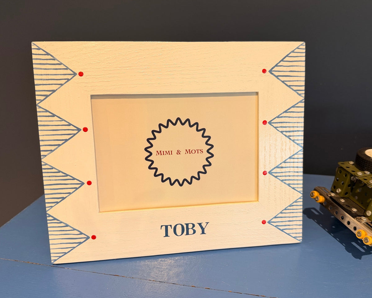 A personalized wooden picture frame with blue geometric patterns, red dots, and the name 'TOBY' at the bottom, displayed on a blue surface next to a toy vehicle.