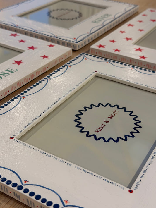 A close-up of a white decorative picture frame with red stars, blue dotted borders, and a central blue scalloped circle containing the text 'Miki & Mavis'.