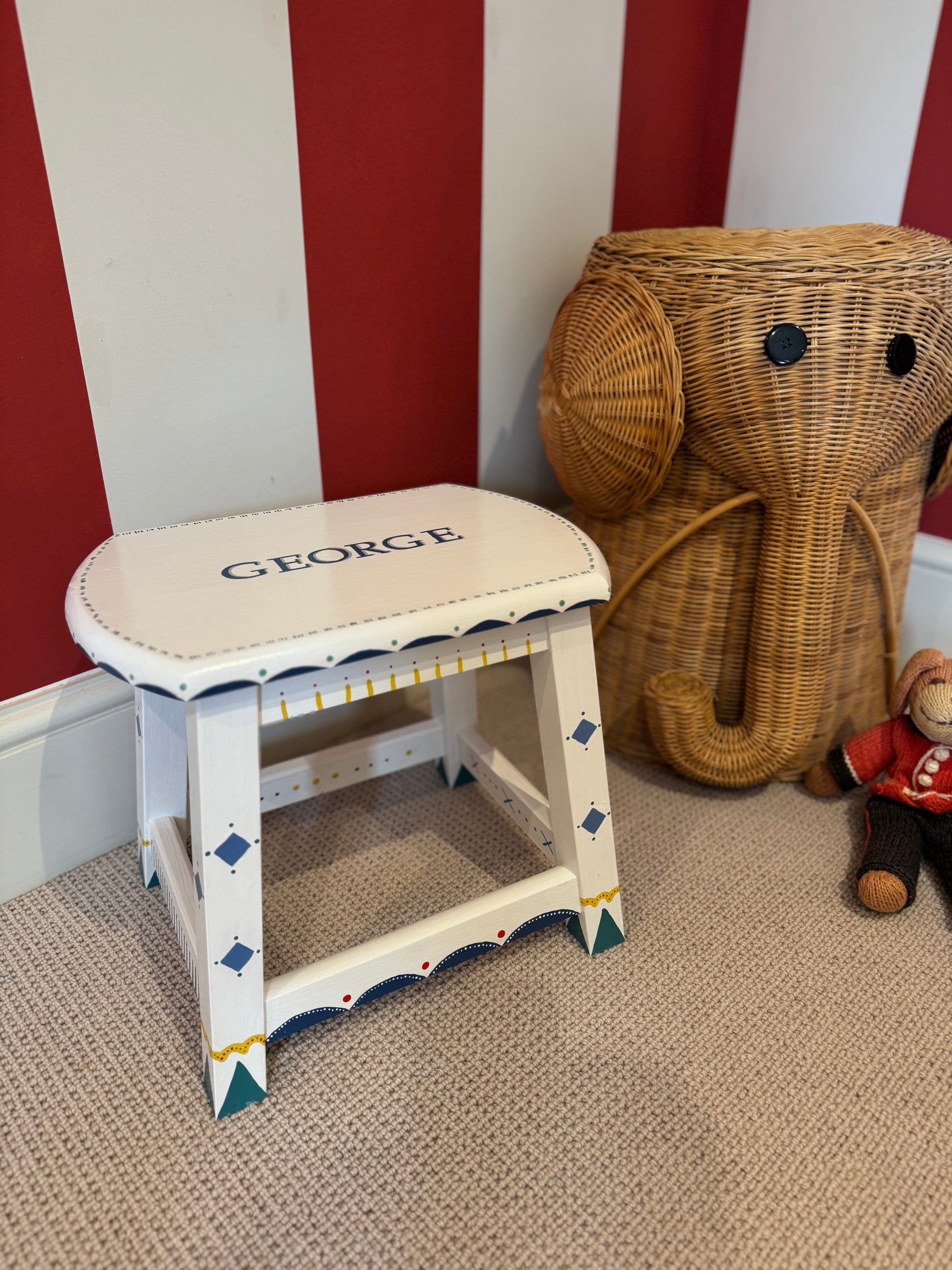 Small white stool with 'GEORGE' on top next to a large wicker elephant toy against a red and white striped wall.