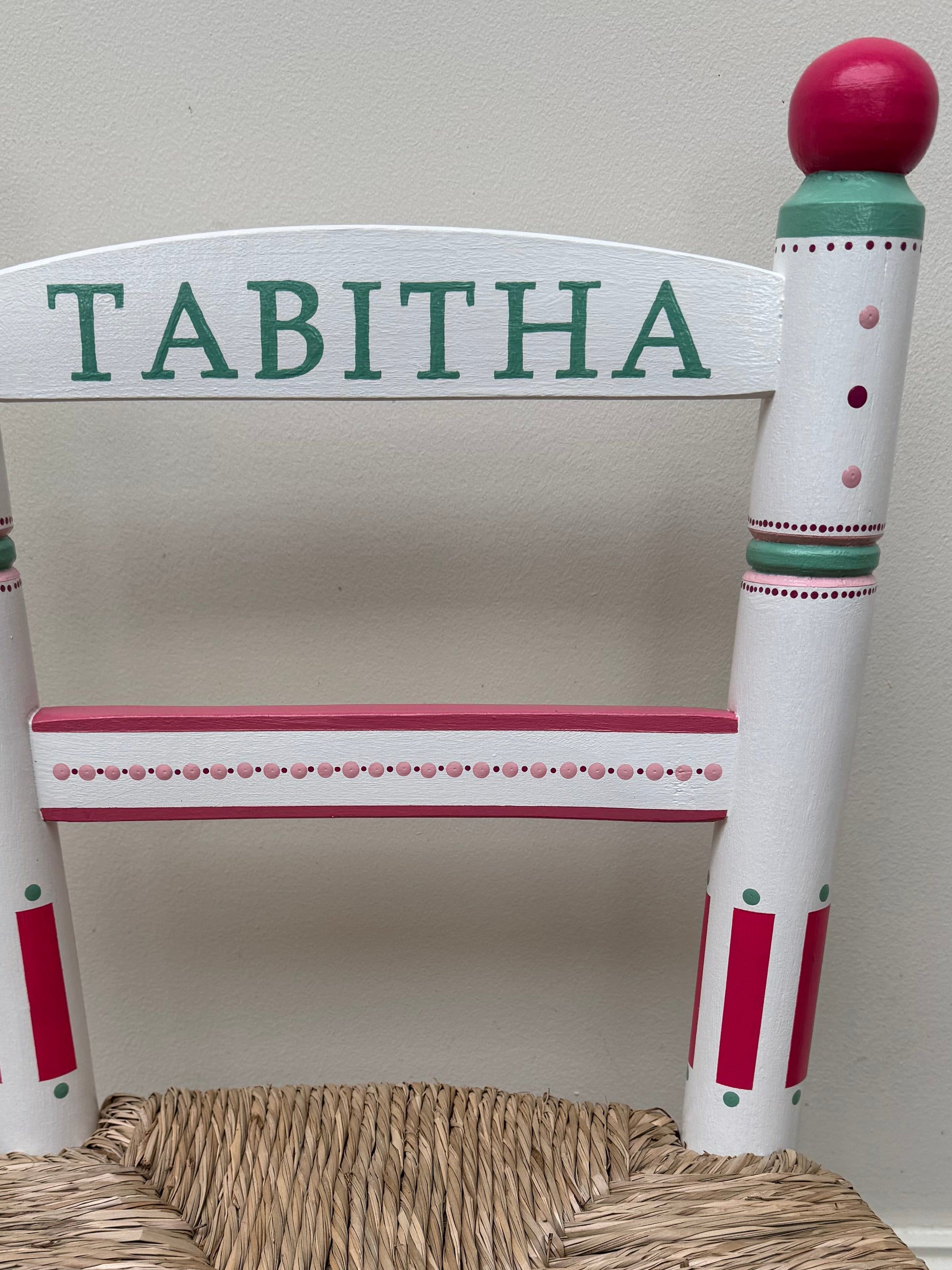 Children's chair with a woven seat and 'Tabitha' on the backrest against a white wall.