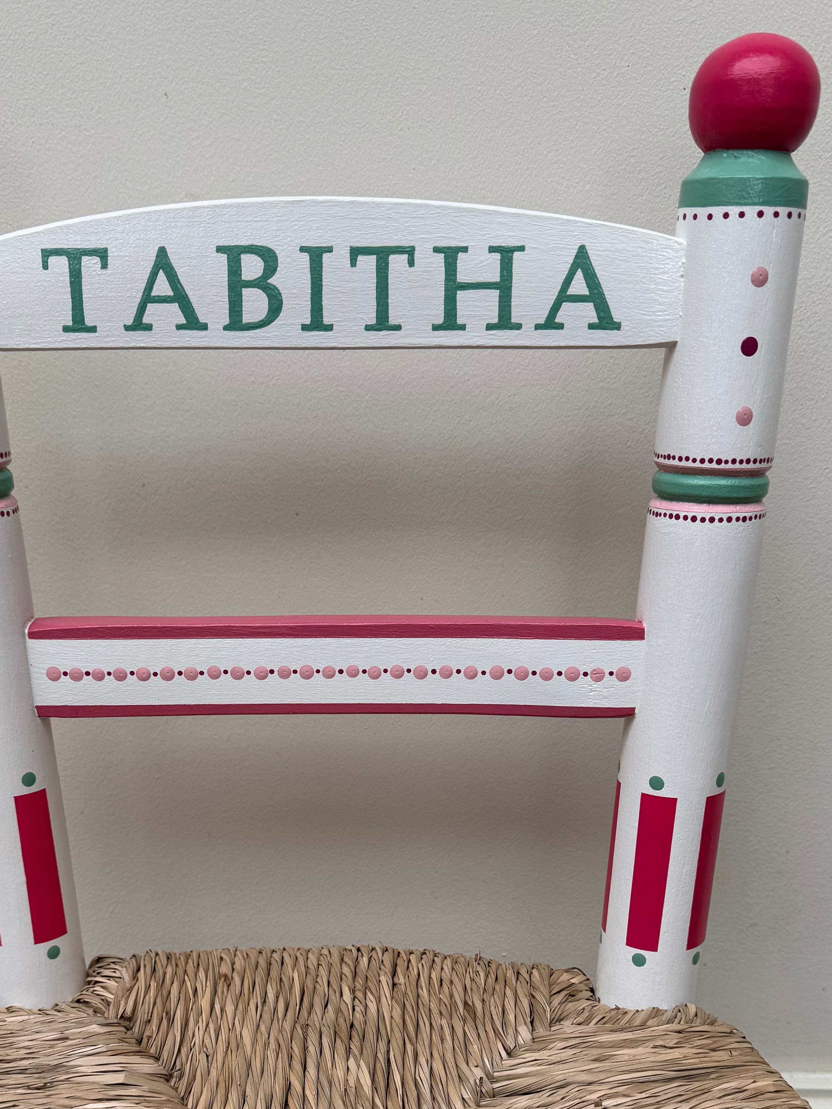 Children's chair with a woven seat and 'Tabitha' on the backrest against a white wall.