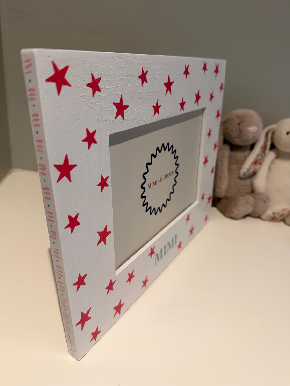 A white wooden picture frame decorated with pink stars and the name 'MIMI' in green, standing on a light surface with plush toys in the background.