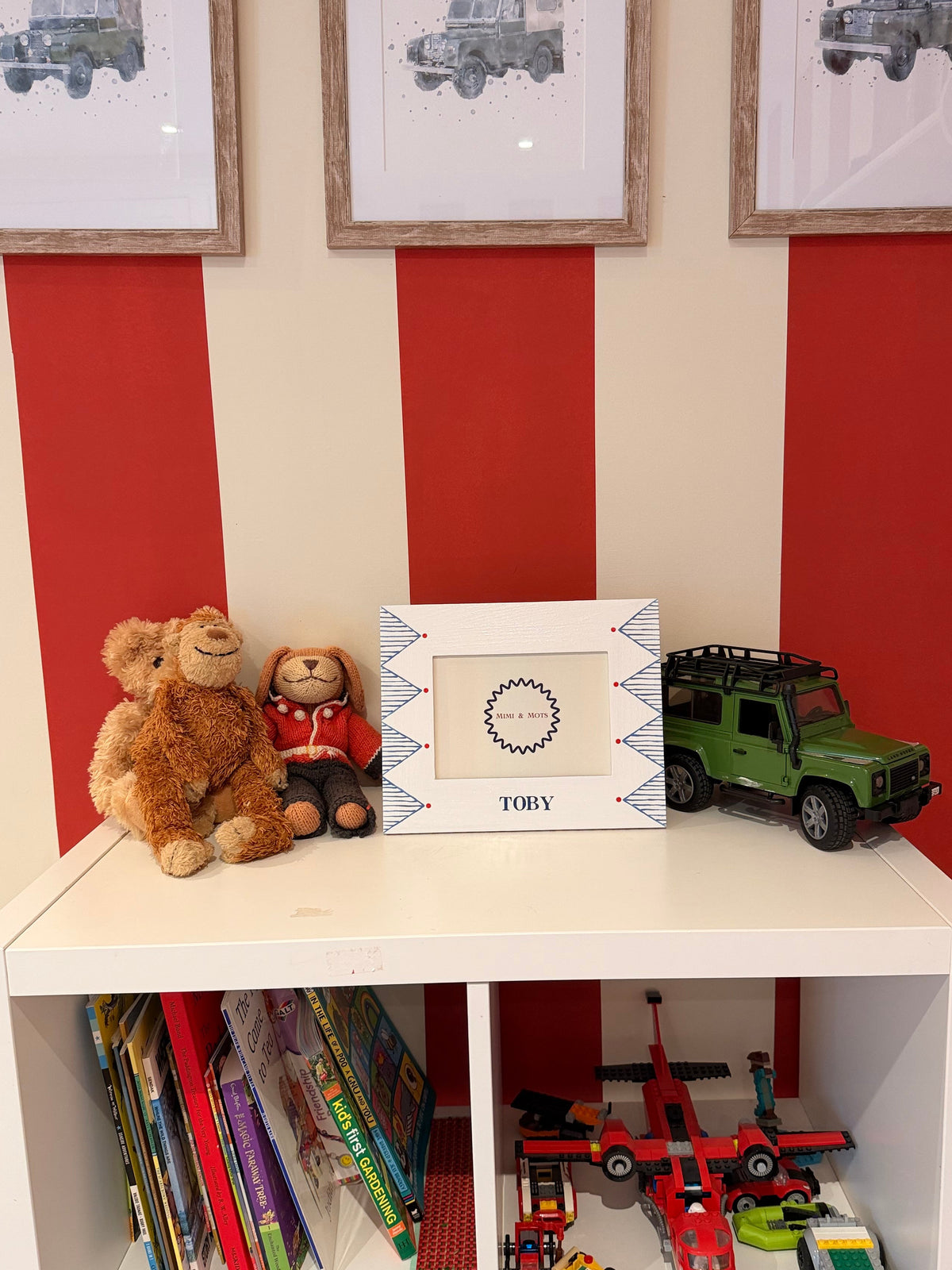 A white children's bookshelf filled with books and toy vehicles, topped with two teddy bears and a framed photo, set against a cream and red striped wall with framed military vehicle prints.